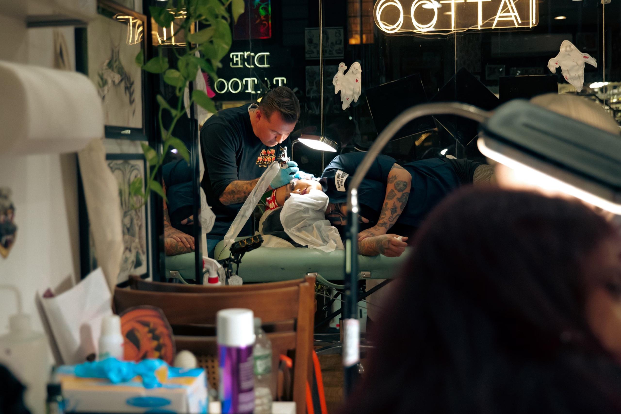Mike Stapleton tattooing a client at night, seen through the shop window under a neon tattoo sign